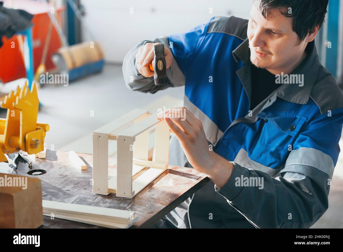 Caucasian carpenter for 30 years in working clothes sits at workbench and makes wooden boxes. Hammers nail with hammer. Authentic workflow scene. View from outside. Stock Photo