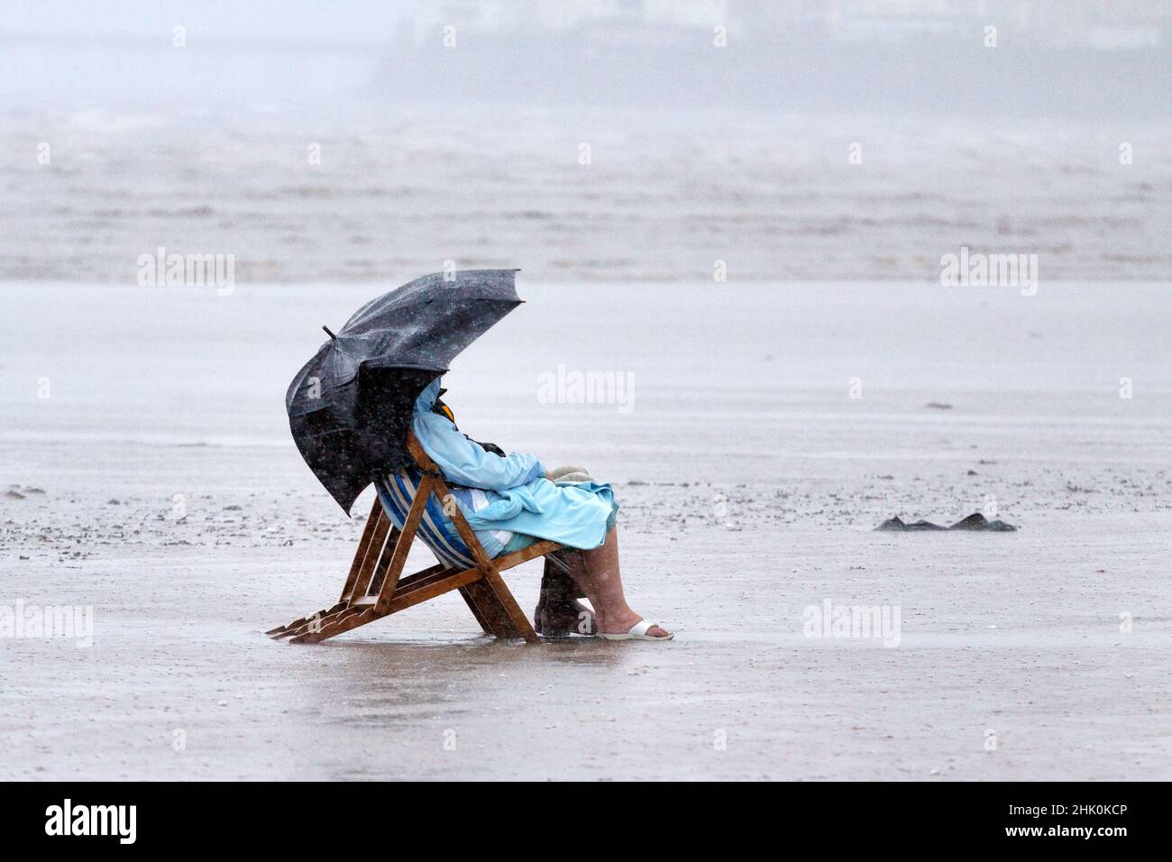A comical scene of a man and a woman sitting in deck chairs. on a British beach, in rain coats during a heavy rain storm inclement summer weather Stock Photo