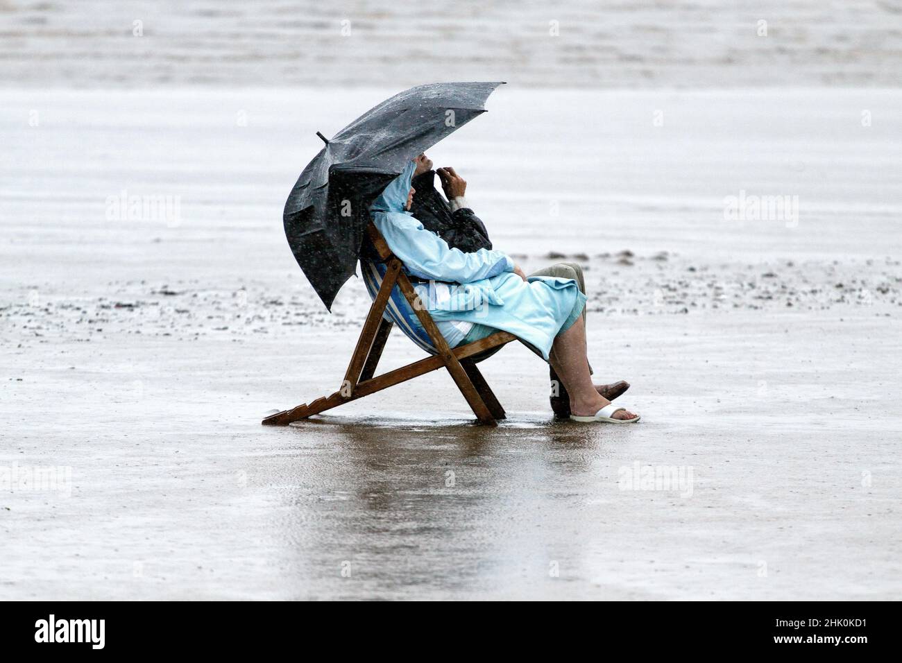 A comical scene of a man and a woman sitting in deck chairs. on a British beach, in rain coats during a heavy rain storm inclement summer weather Stock Photo