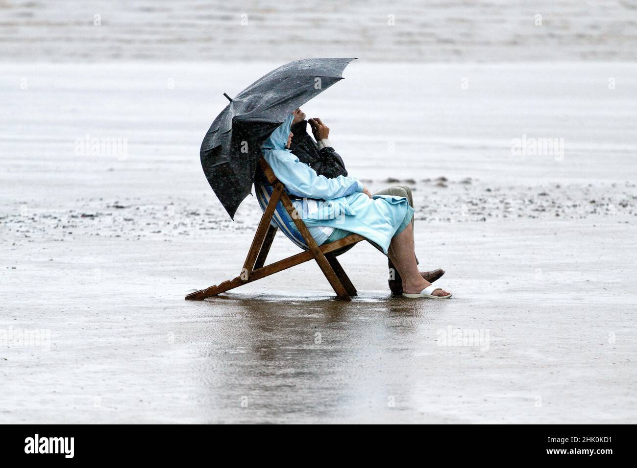 A wet weather scene of a man and a woman sitting in deck chairs. on a British beach, in rain coats during a heavy rain storm during summer weather Stock Photo