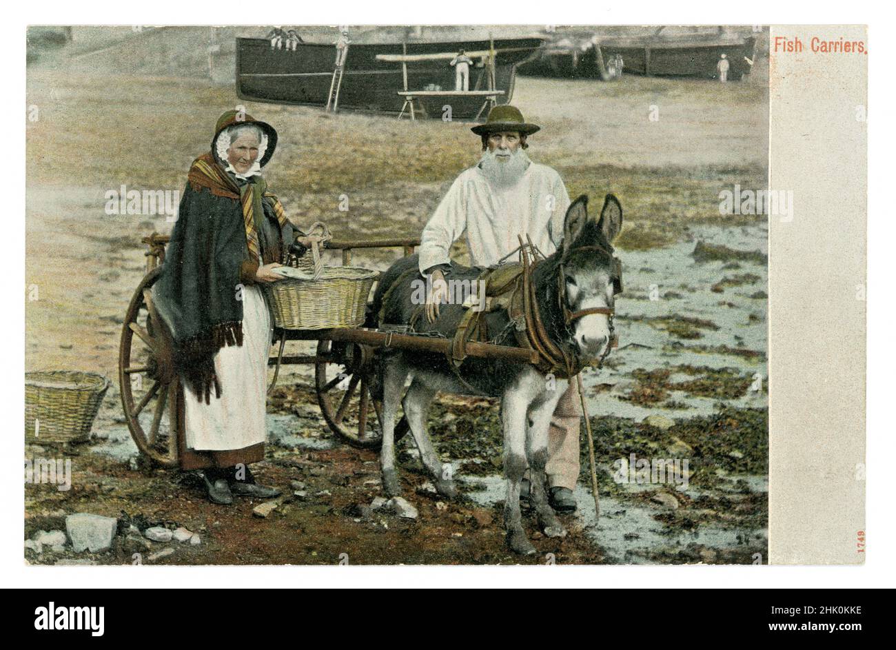 Original Edwardian era tinted greetings souvenir postcard of Cornish Fish Carriers, fishwife, with donkey cart on the waterfront, Newlyn harbour, Cornwall, U.K. The man is a local called Billy Renfree and his fishwife is filling up her basket with fish. She wears a traditional shawl and apron known as ‘towsers’, dated to 1906 from a posted postcard. Stock Photo