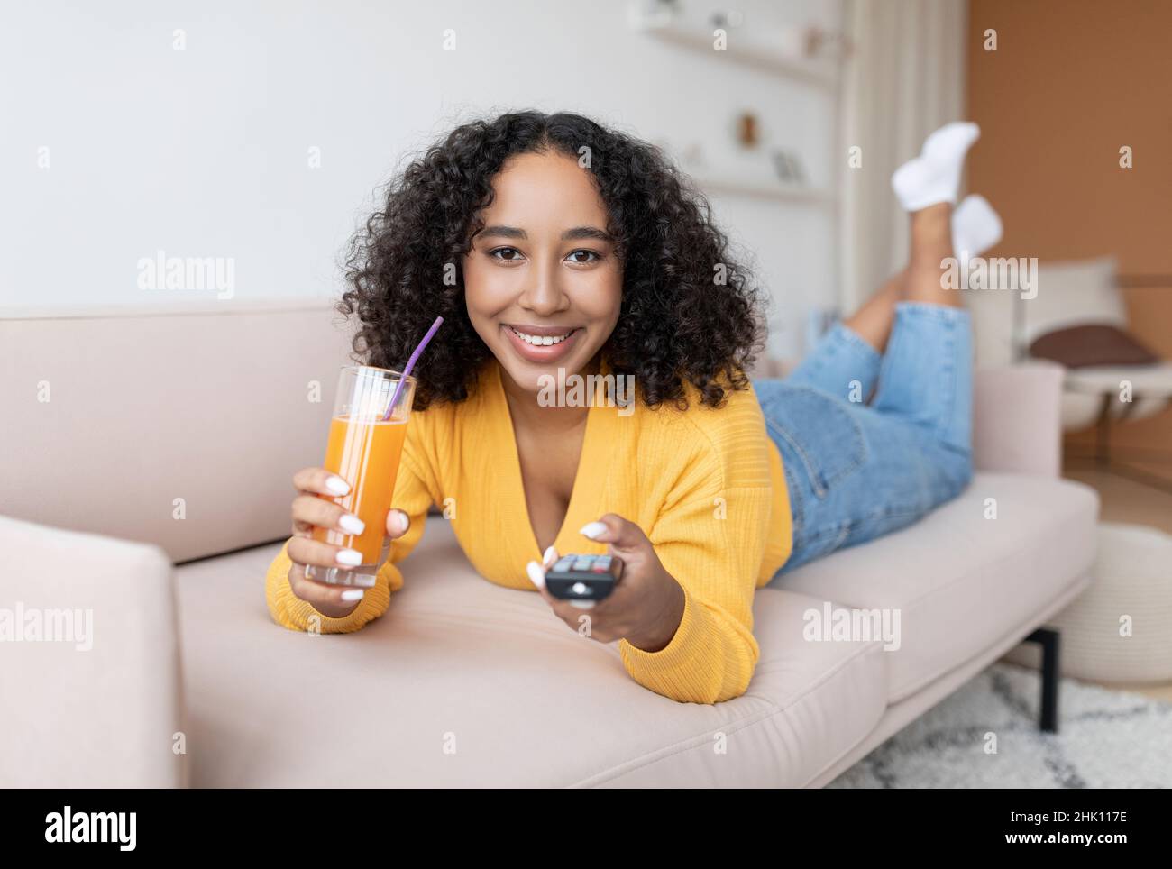 Happy young black lady lying on couch with TV remote control and glass of orange juice, switching channels at home Stock Photo