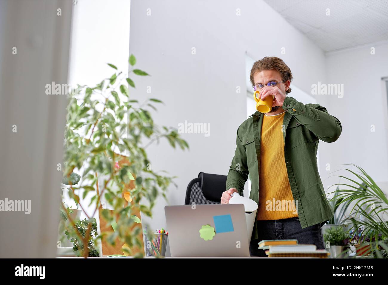 young pleasant guy stand behind office desk drinking tea, break time at work place. handsome caucasian man in casual wear have rest, plants in office. Stock Photo