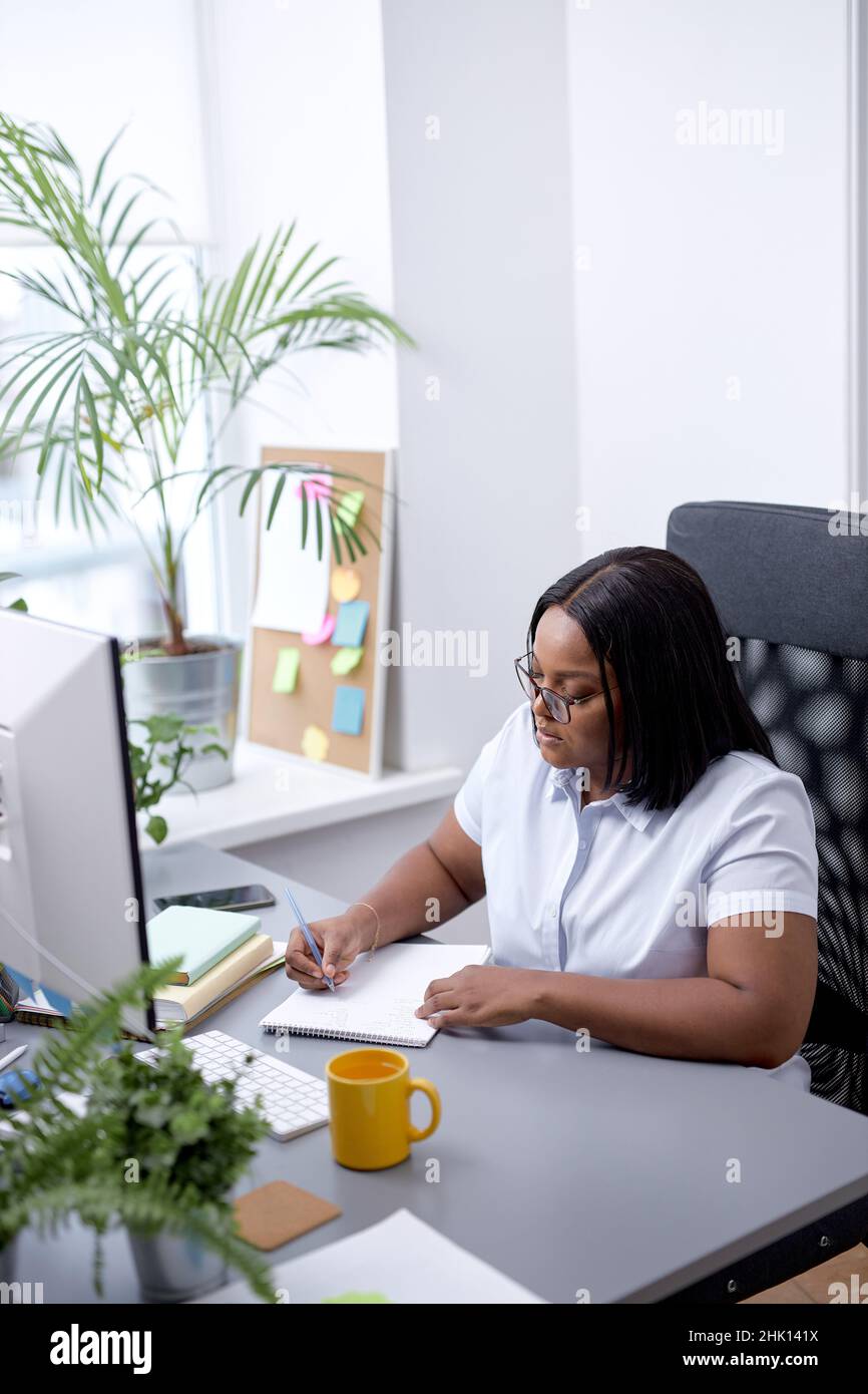 Black Office Woman Sitting At Desk Working On Laptop Taking Notes At Work Indoor. Business Schedule Concept. Beautiful Lady in White Shirt Concentrate Stock Photo