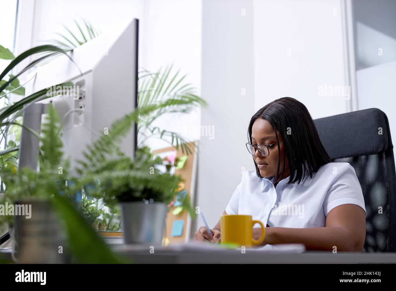 Black lady office worker concentrated on work, writing documents information, sit alone in bright cozy modern office, use pc computer, dressed in form Stock Photo