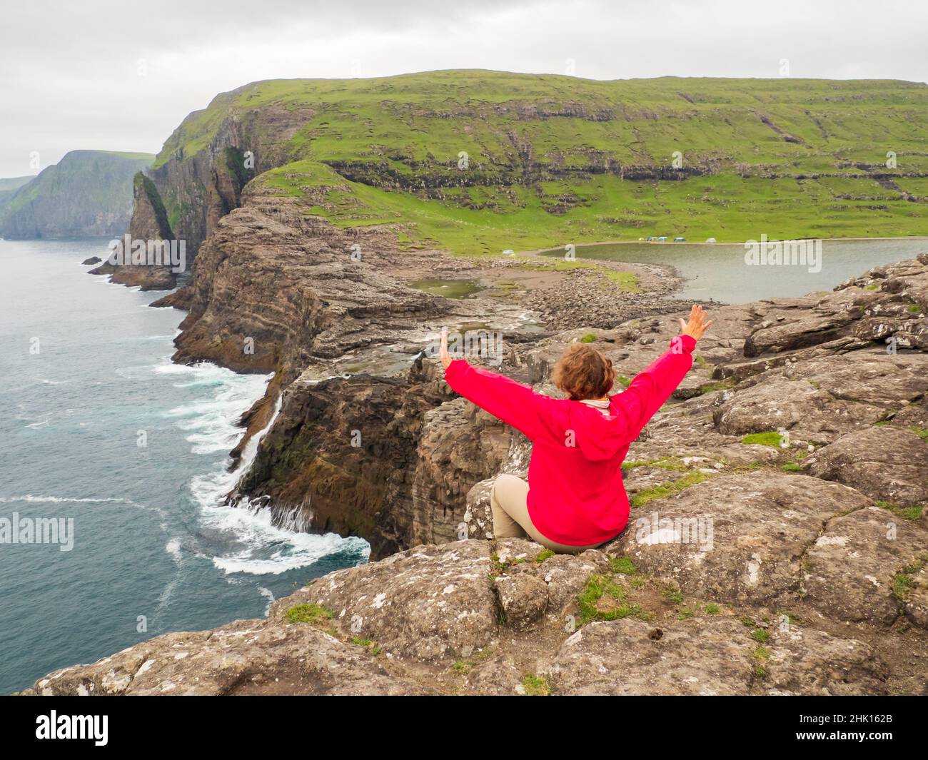 A tourist in a red jacket sits on a rocky shore above the Bøsdalafossur Waterfall, which flows from Lake Sørvágsvatn / Leitisvatn into the Atlantic Oc Stock Photo