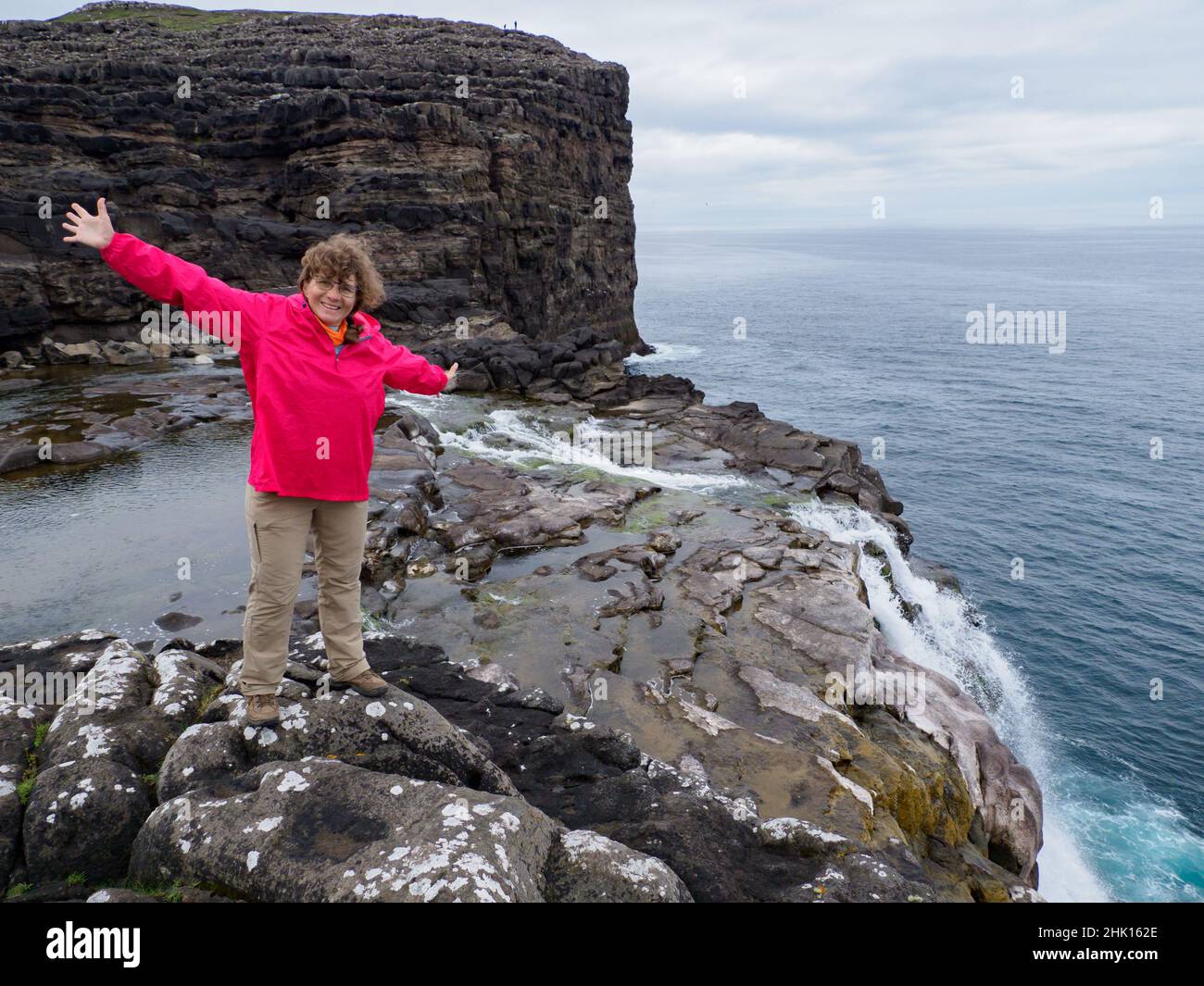 A tourist in a red jacket sits on a rocky shore above the Bøsdalafossur Waterfall, which flows from Lake Sørvágsvatn / Leitisvatn into the Atlantic Oc Stock Photo