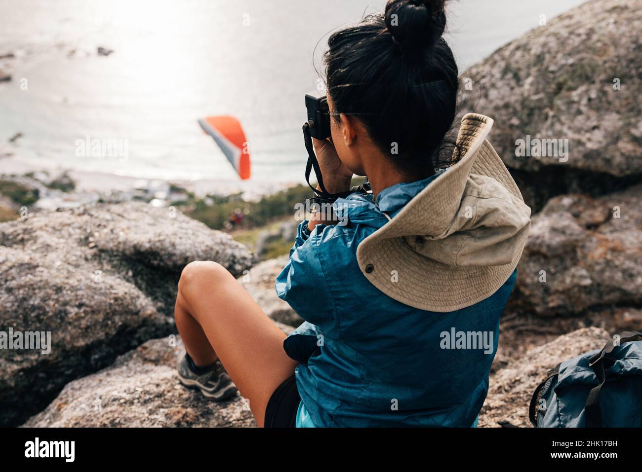 Woman sitting on the cliff making photographs on a film camera. Back view of female trekker resting on a mountain top with a camera. Stock Photo