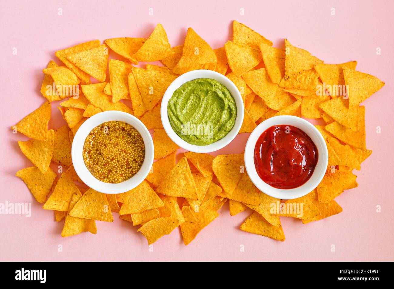 Heap of corn tortilla chips with french mustard , guacamole and ketchup on a pink background. Top ivew. Stock Photo