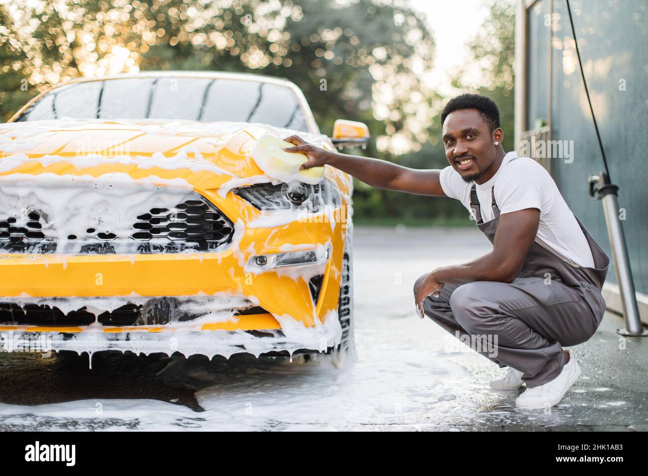 slids udskiftelig Whitney Car detailing wash at outdoors car wash service. Handsome African American  man in white t-shirt and overalls washing car radiator grille and hood of  his new yellow sport car with foam and