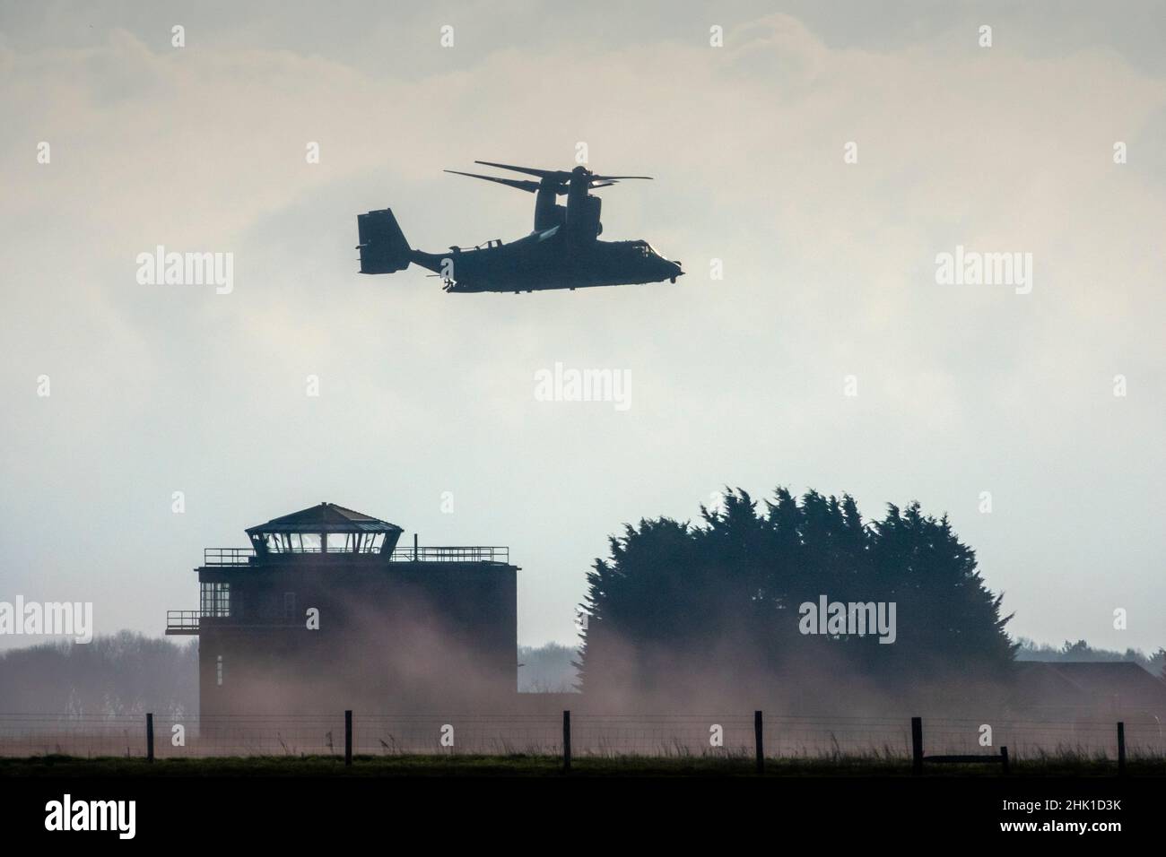 A partly silhouetted distance side shot of a Bell Boeing V-22 Osprey hovering over an airfield control tower sucking up local dust Stock Photo