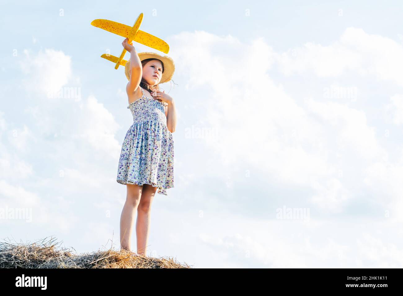 Portrait of little girl in hat and dress playing flying yellow toy airplane, standing on haystack. Taking aim in sky. Light sunny day. Having adventur Stock Photo