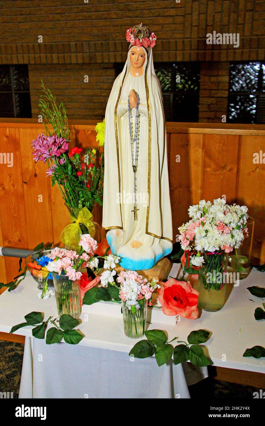 Statue of Our Lady of Fatima holding rosary beads and wearing a crown of flowers on her crown. She is sitting on a table surrounded by cut flowers in Stock Photo