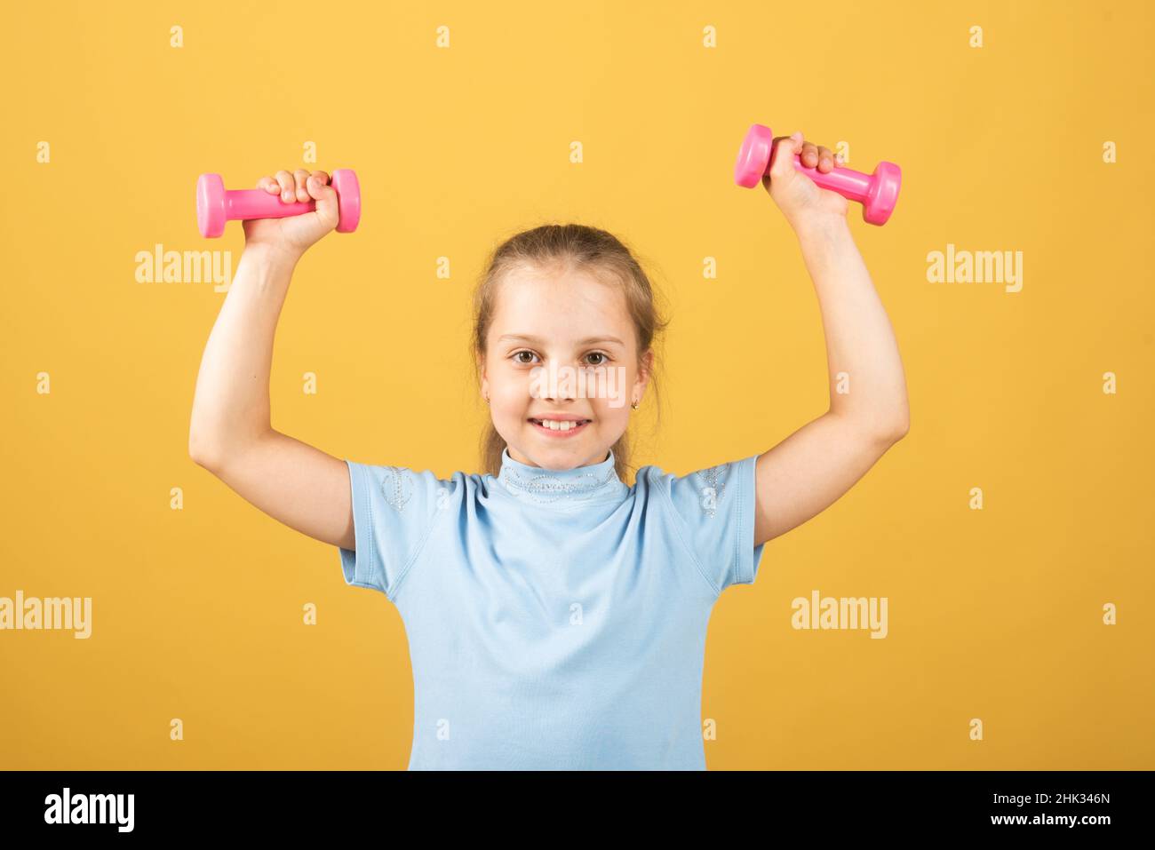 Cute little girl doing exercises with dumbbells in yellow background ...