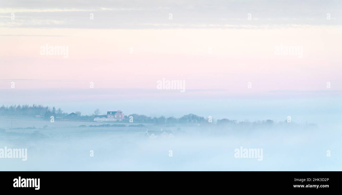 A lonely house stands above a mist shrouded landscape during a pastel sunrise in the Lower Wharfe Valley, viewed from Almscliffe Crag. Stock Photo