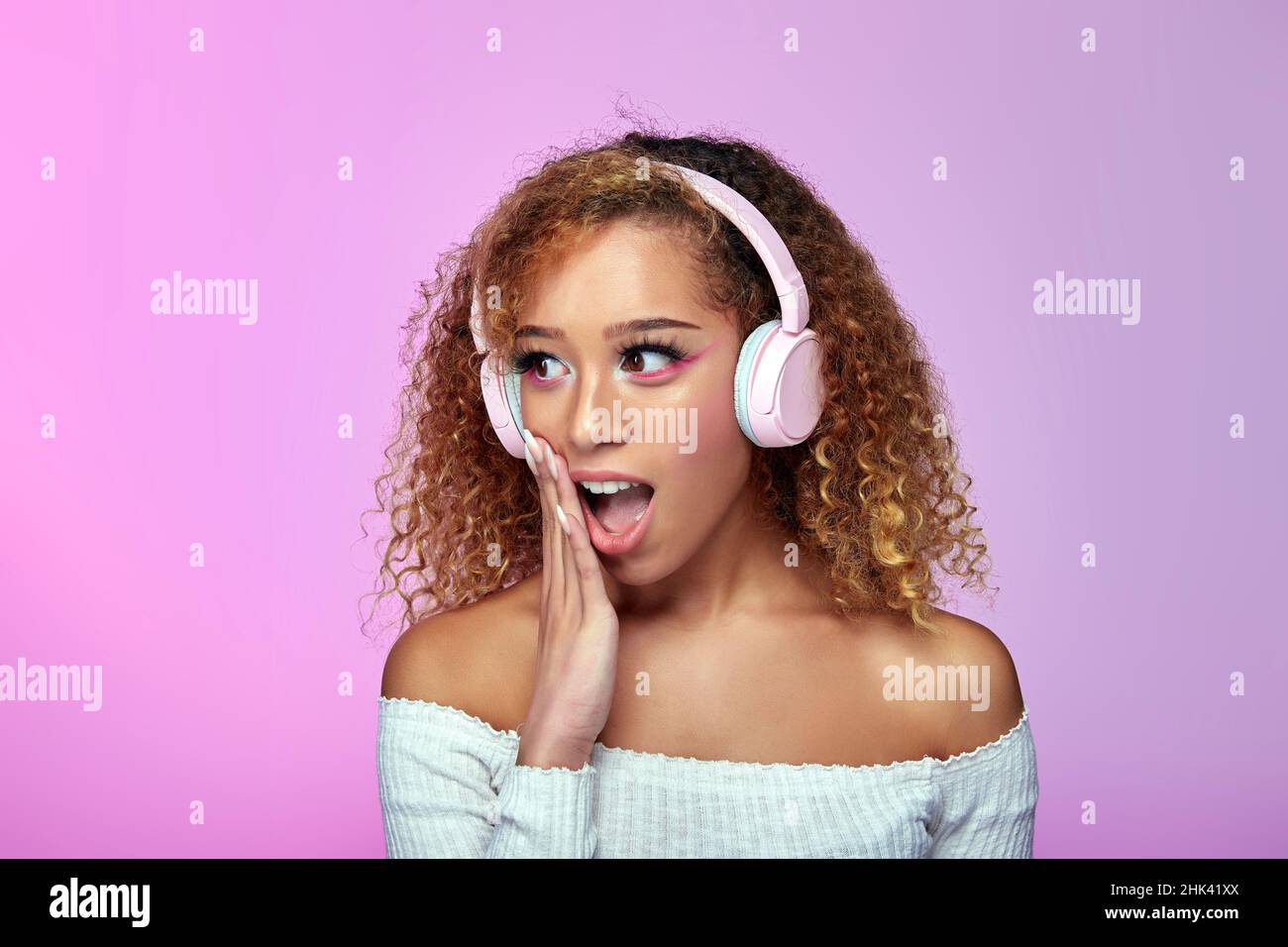 Shocked African American curly haired female listening to songs in headphones while expressing astonishment with opened mouth on gradient pink background in studio Stock Photo