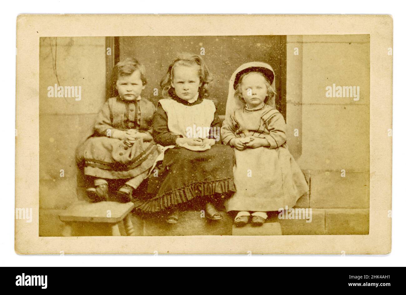 Original Victorian Carte de Visite (CDV) or visiting card, of 3 young children sitting on a step outside a large house, each holds an apple. All are wearing dresses. The child on the left appears to be a boy - it was normal to dress young boys in girls clothes in Victorian times. circa 1860's, U.K. Stock Photo