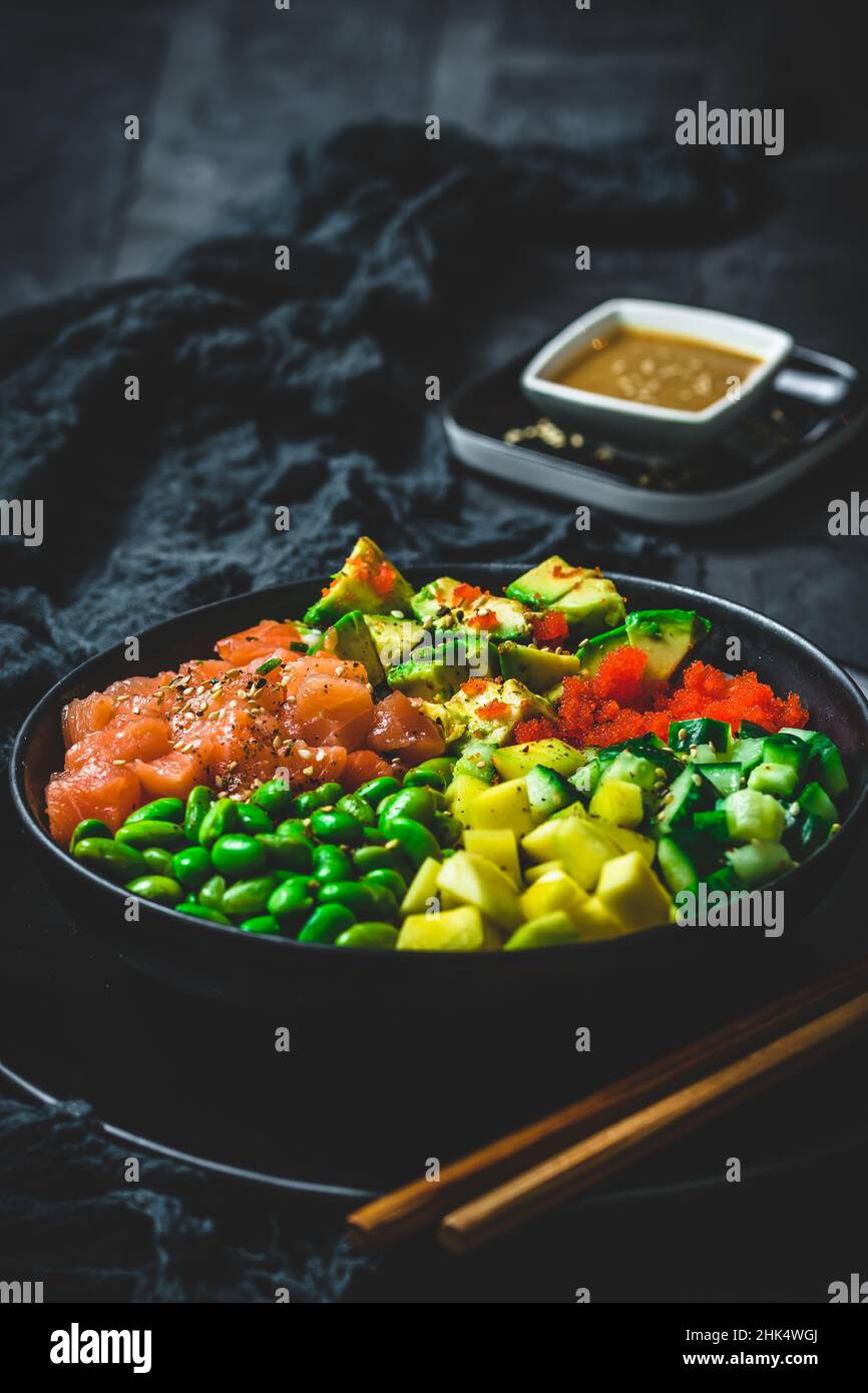 Japanese poke or buddha bowl with salmon, avocado, roe, mango, pies and cucumber in a black bowl on black background, vertical Stock Photo