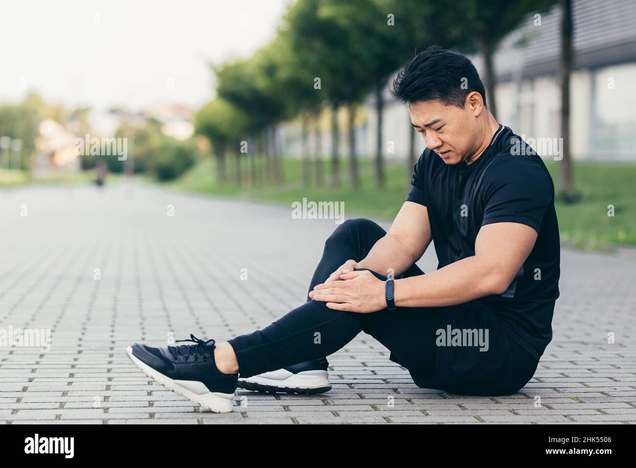 Asian man after fitness workout and jogging sits on the ground and suffers from leg pain, massages leg muscles Stock Photo