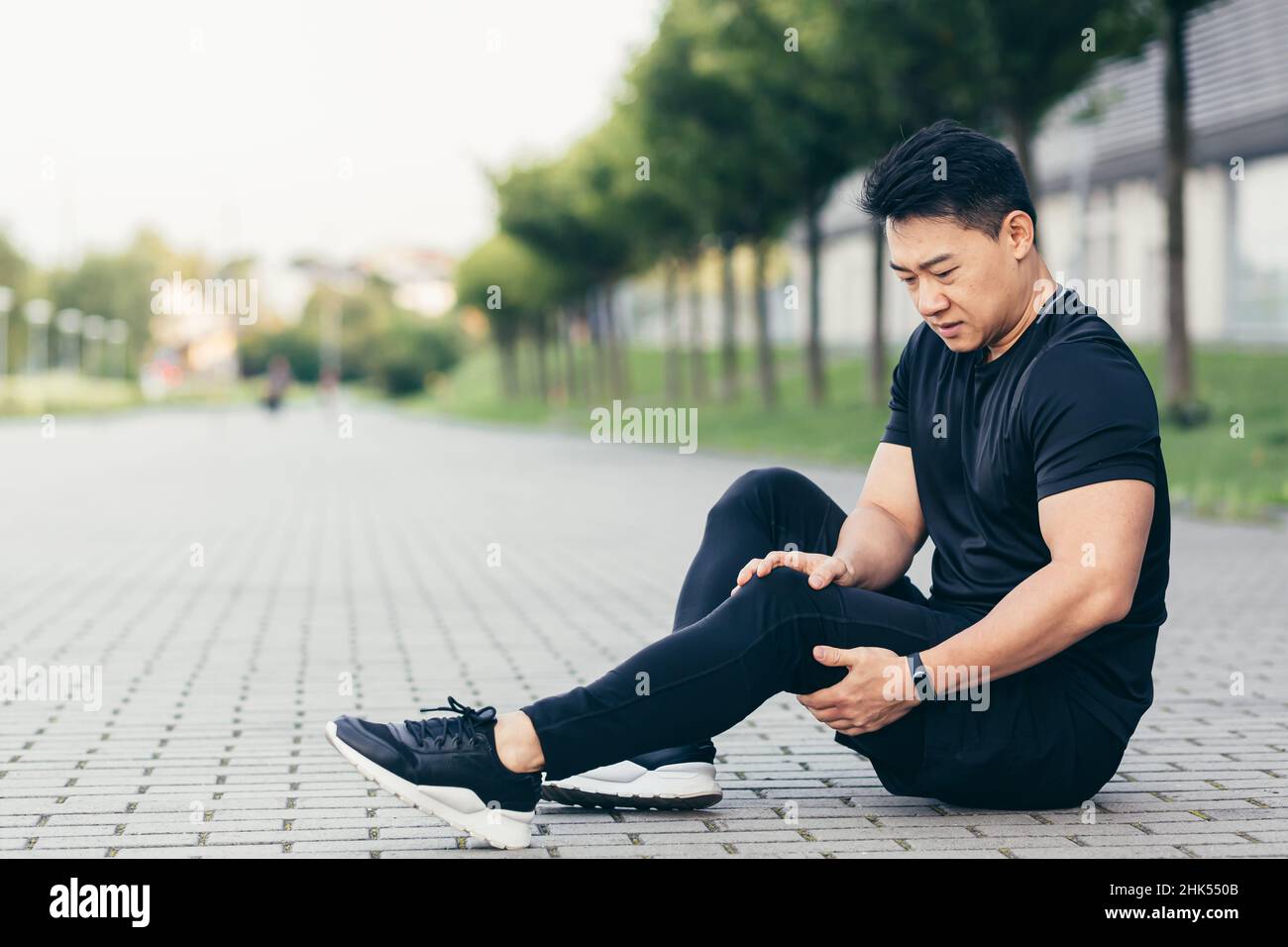 Asian man after fitness workout and jogging sits on the ground and suffers from leg pain, massages leg muscles Stock Photo