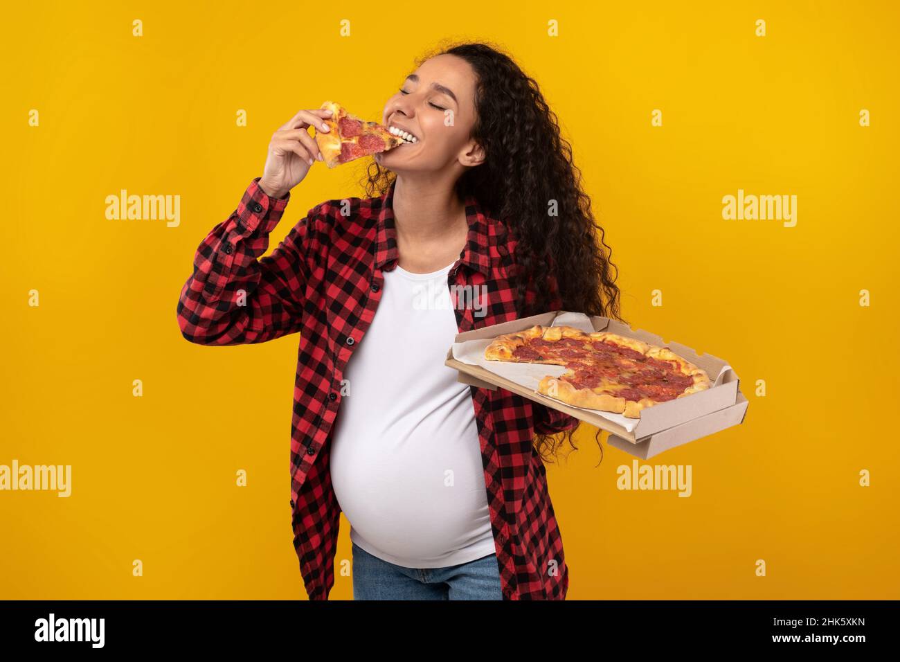 Happy Pregnant Latin Lady Holding Box Biting Pizza At Studio Stock Photo