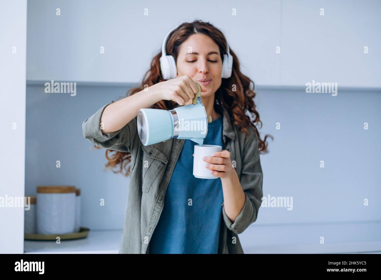 Curly-haired young woman with loose hairstyle dances to loud music in wireless headphones with coffee in cup and coffeepot Stock Photo