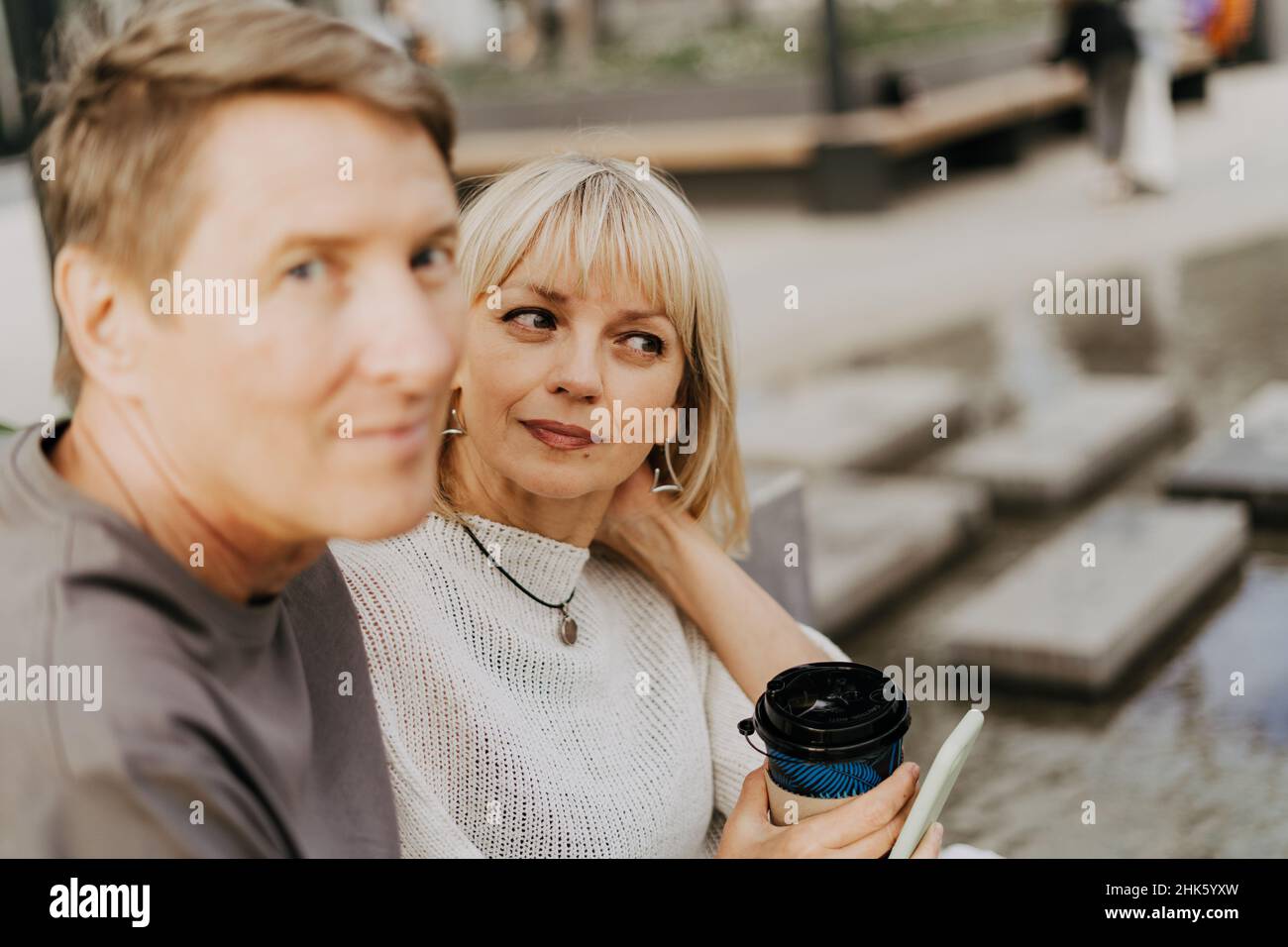 An adult mature happy couple in love sitting on bench outdoors in city street park. A blonde caucasian man and woman spend time together and drinking Stock Photo