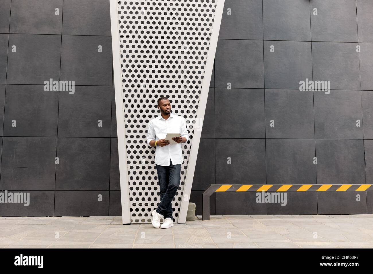 Confident serious businessman hold tablet, standing near the office, lost in thoughts about new startup. Stylish african american male employee in Stock Photo