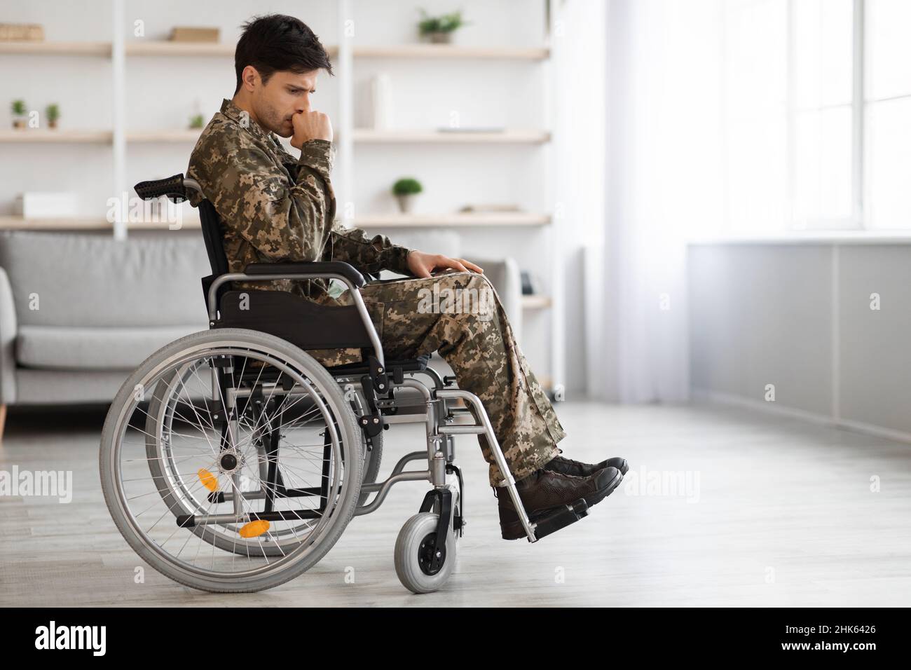 Upset young male looking at camera with sad look. He is sitting in invalid chairing wearing military uniform. Isolated on grey background. Copy space Stock Photo