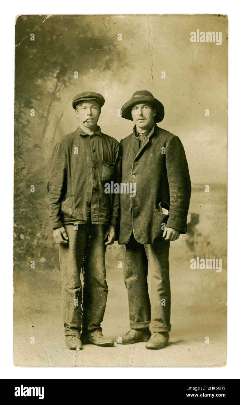 Original, clear, Edwardian era postcard of 2 poor working class men, friends, characters, possibly itinerant labourers, also known as navvies. The men are wearing dirty workwear, jackets, hat and flat cap, one smoking a cigar (he looks like he could be trouble, a rogue!), the other is very shabby looking wearing a jacket with the buttons undone and a crumpled hat who seems to have his lunch in his pocket. They are photographed in their scruffy working clothes as if they have been hurriedly photographed after working outside.  U.K. circa 1905. Stock Photo