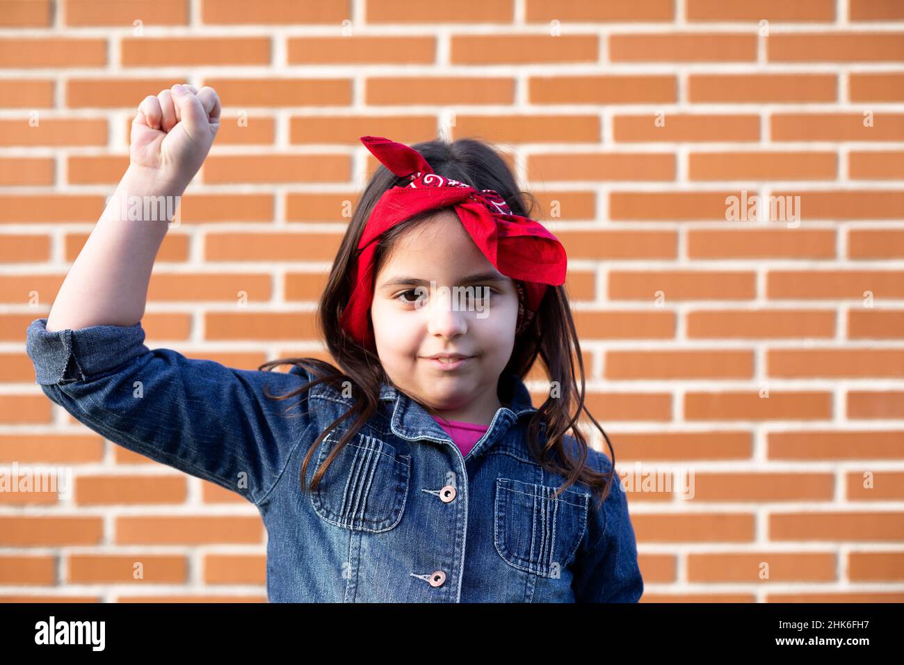 Little girl in representative Women's Day dress with raised fist. Background of brick wall. Space for text. Stock Photo