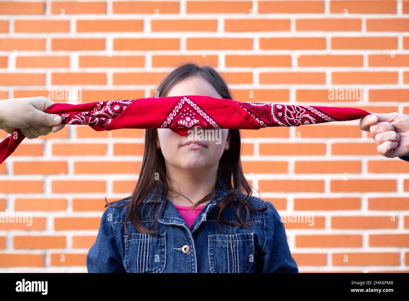 Portrait of a little girl with her eyes covered by a scarf isolated on a brick wall. Concept of innocence and justice. Stock Photo
