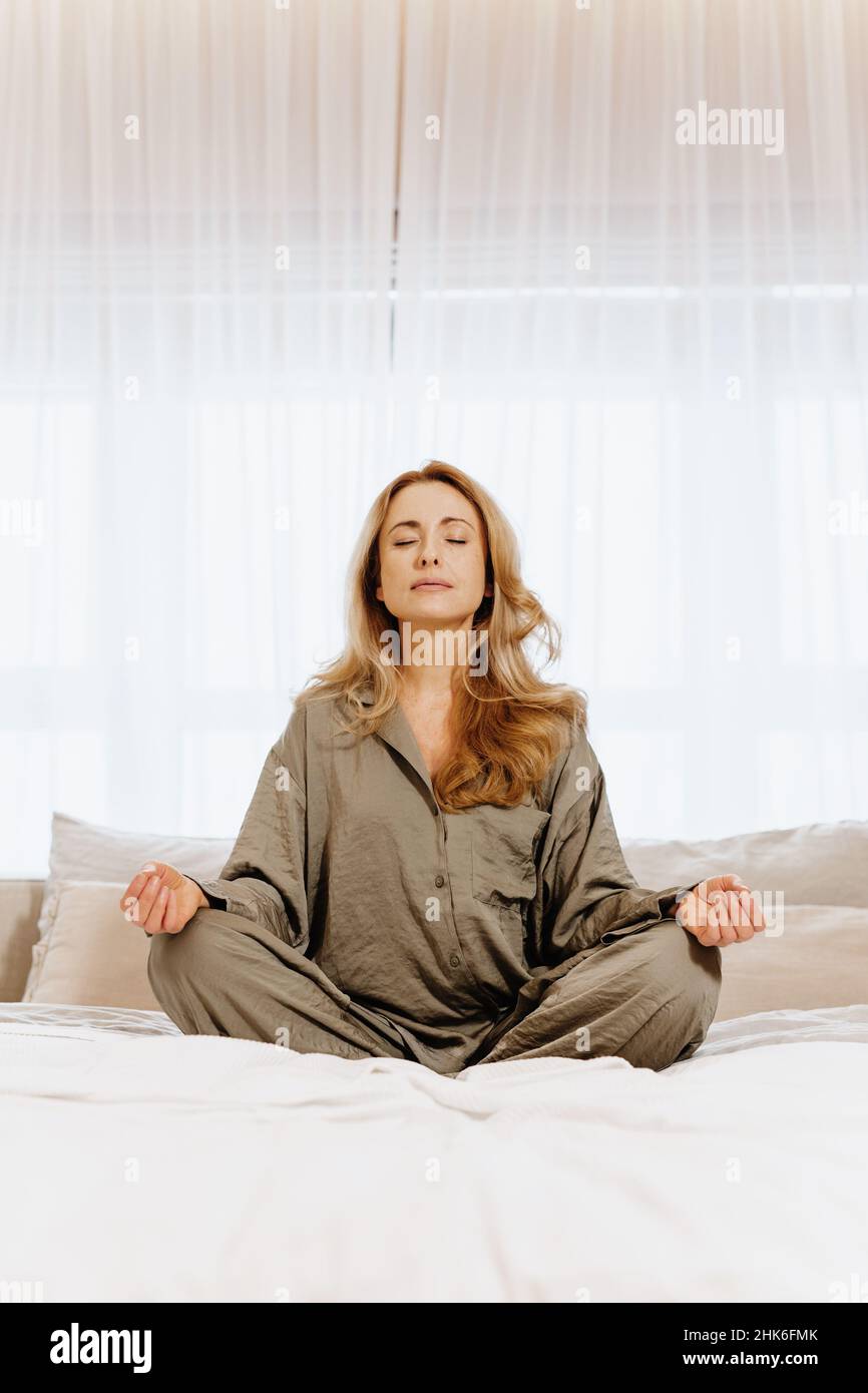 Adult woman with closed eyes in sleepwear meditating on bed in bedroom. Stock Photo