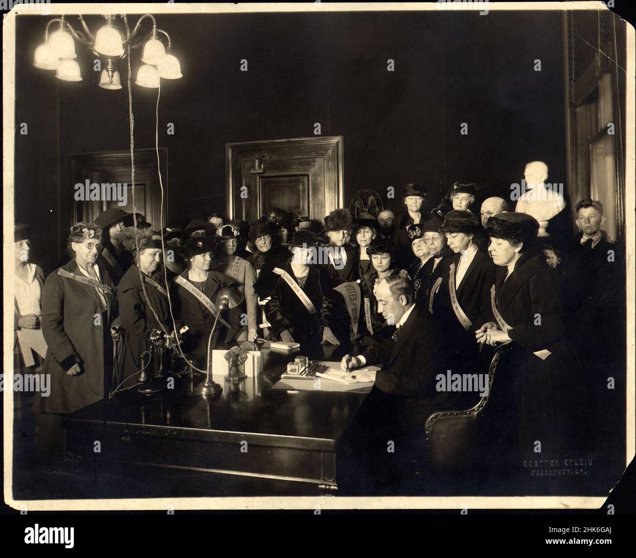 Governor Edwin P. Morrow signing the Anthony Amendment--Ky. was the twenty-fourth state to ratify, January 6, 1920. Large group, mostly women wearing 'Votes for Women' banners, standing around desk in office as Governor Edwin Morrow signs amendment. Stock Photo