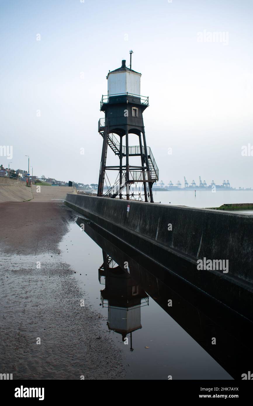 Dovercourt Lighthouse is reflected in a puddle on the promenade on a cloudy and misty day.  Port of Felixstowe can be seen in the background. Stock Photo
