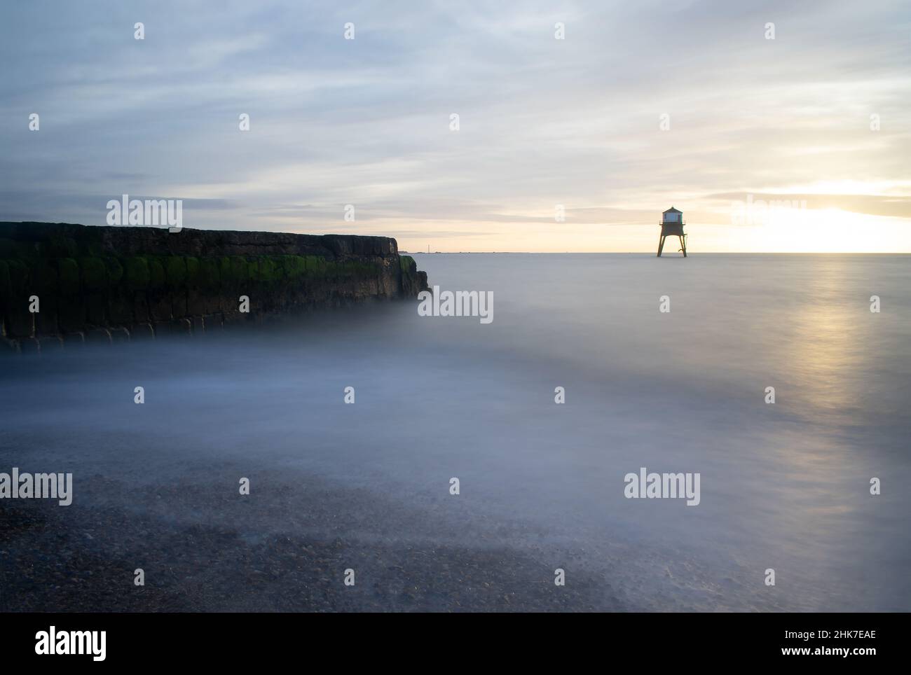 Waves break across the shingle beach and mossy breakwater during sunrise at Dovercourt Lighthouse Harwich & Dovercourt, North Essex, England. Stock Photo