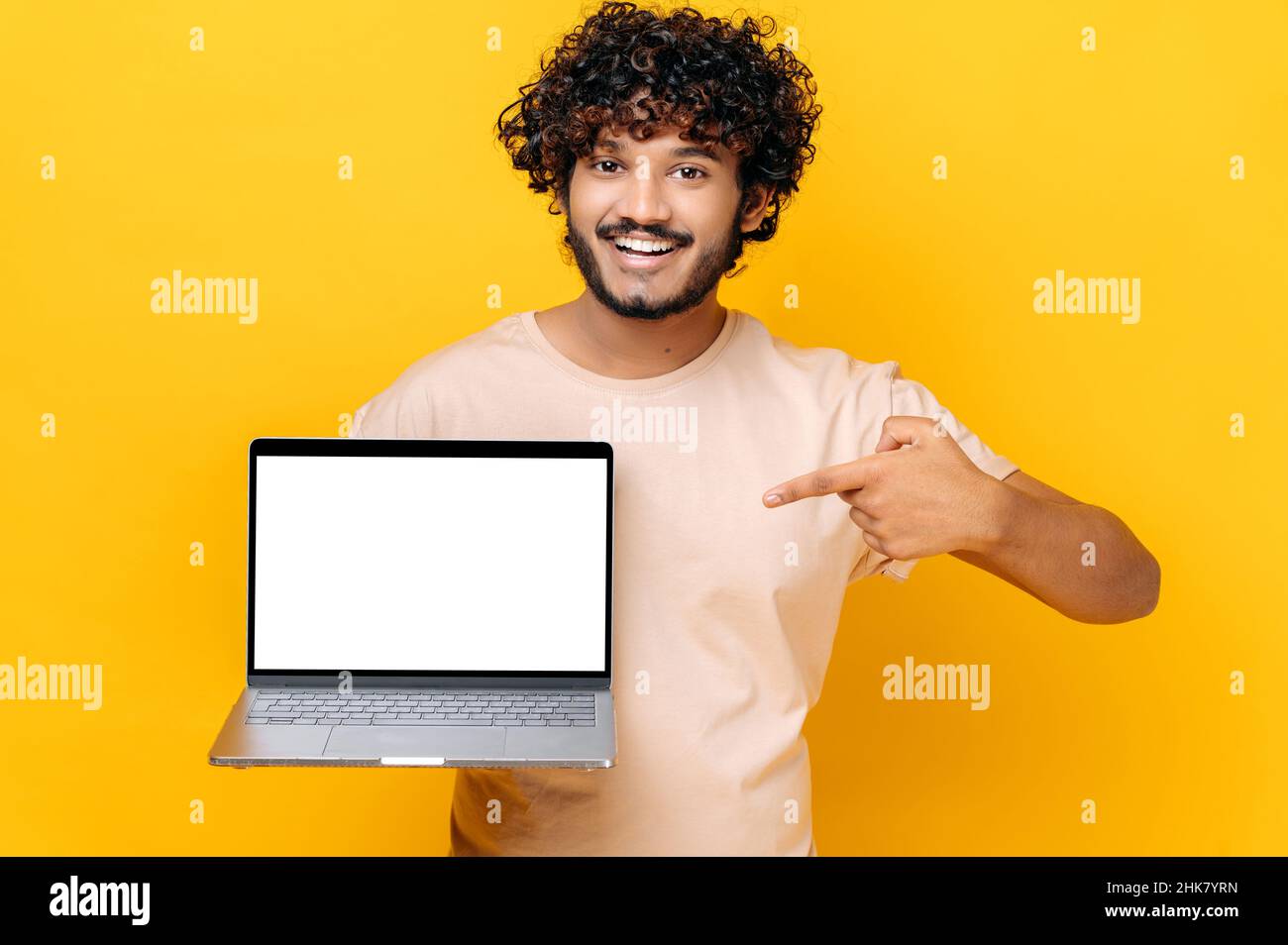 Positive confident Indian stylish curly guy, holds an open laptop with blank white screen, points finger at it, standing on isolated orange background, smiling friendly. Copy space, mock-up concept Stock Photo