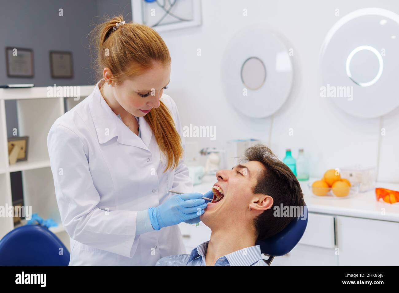 Redhead woman in white robe and latex gloves with ginger ponytail using mirror to inspect teeth of man with opened mouth in dental clinic Stock Photo