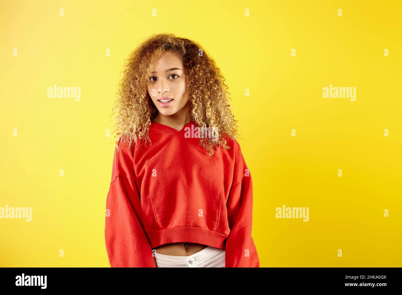 Confident young African American woman in stylish red hoodie and eyeglasses standing on yellow background in studio  Stock Photo