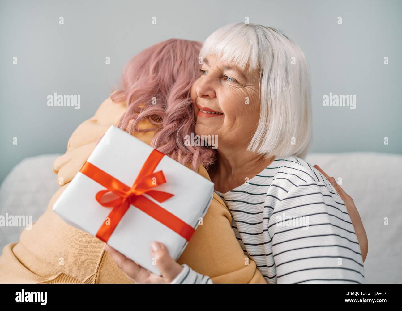 International Women Day. Beautiful happy caucasian daughter giving mother gift box for mothers day sitting on sofa at home Stock Photo