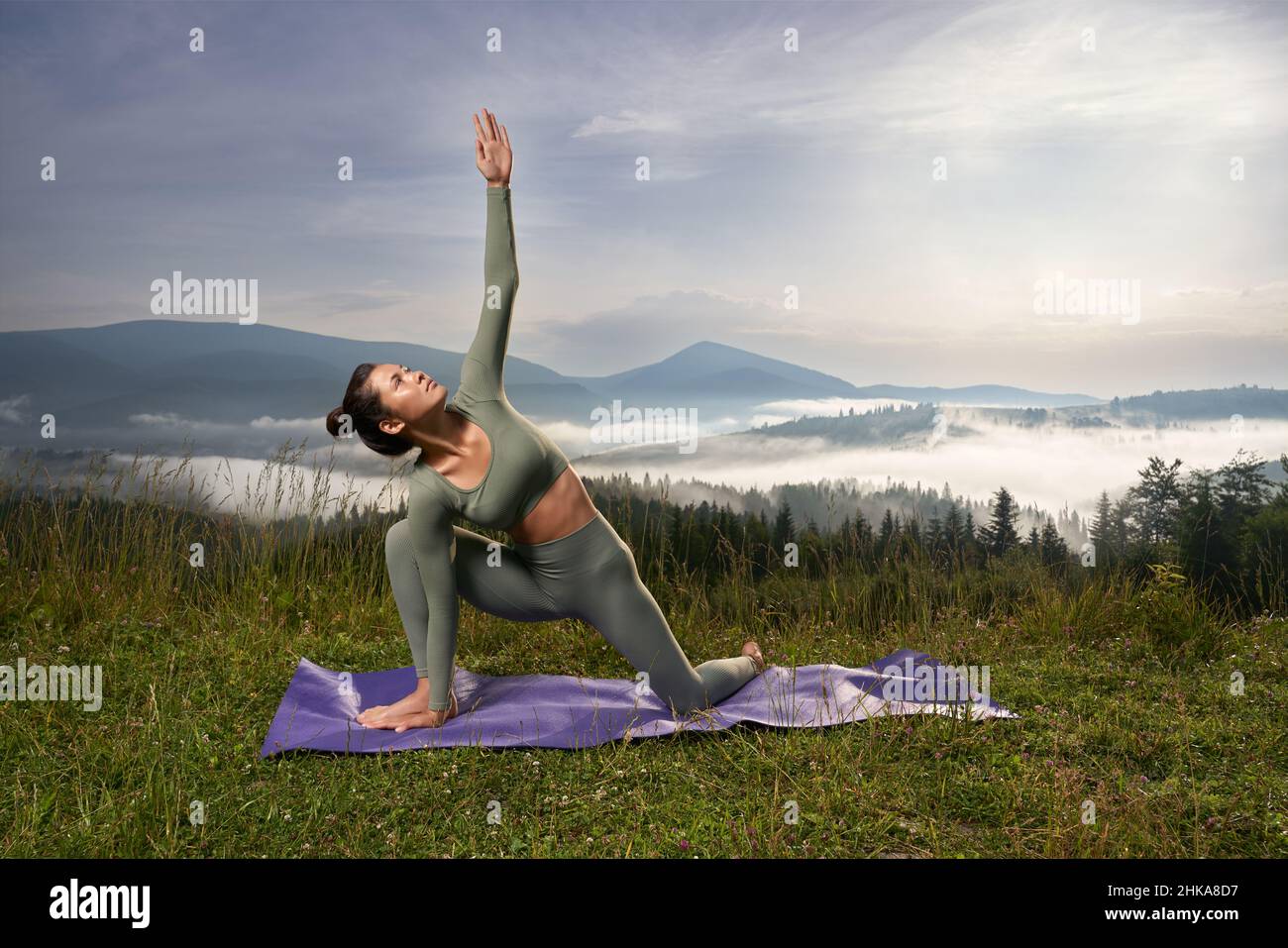 Brunette woman looking up doing Utthita Trikonasana on knees among mountains nature. Pretty relaxing woman have practice of yoga at sunrise time, deeply breathing. Concept of yoga. Stock Photo
