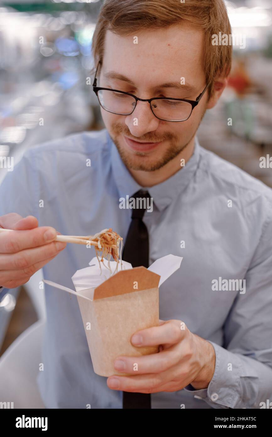 Smiling businessman in glasses eating chinese wok from box on food court. Lunch time. Stock Photo