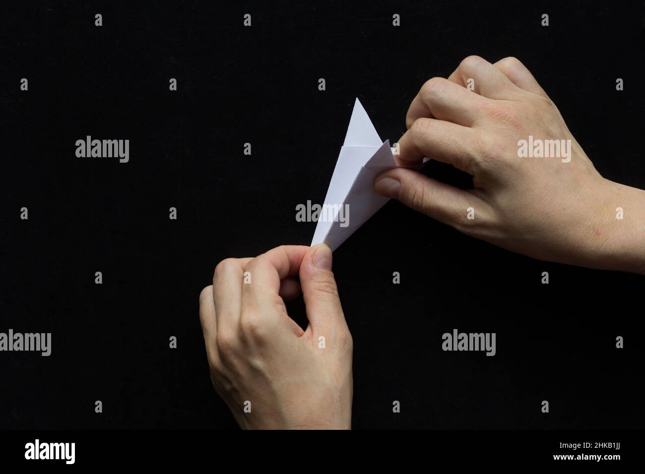 Making snowflake. Step 8. Woman hands folding in half paper triangle with folded one third parts over on black background Stock Photo