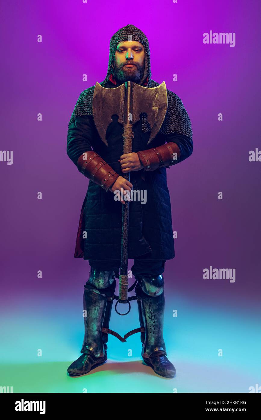 Studio shot of young bearded man in image of medieval warrior, archery in chain armor isolated over purple background in neon. Comparison of eras Stock Photo