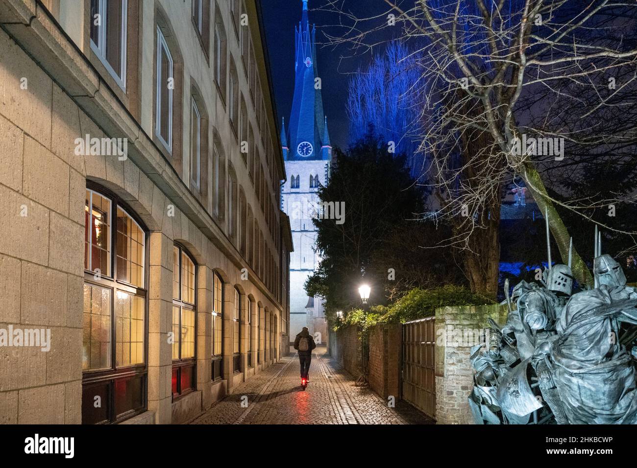 Man riding an electric scooter through Müller-Schlösser-Gasse with city elevation monument and St.Lambertus church at night in the old town of Düsseldorf, NRW, Germany on 11.12.2021 Stock Photo