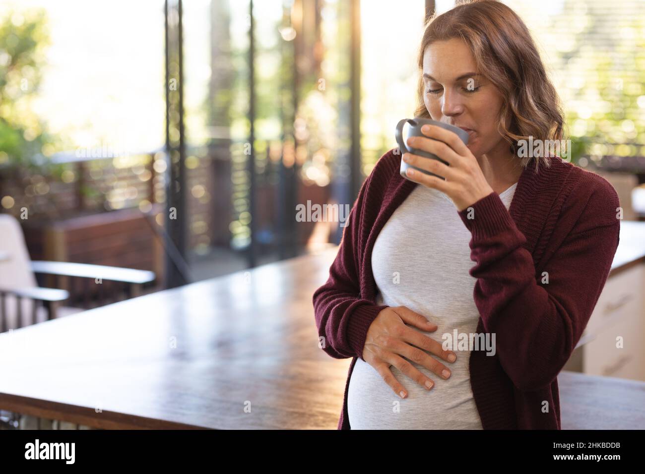 Caucasian pregnant woman touching her belly and drinking coffee at home Stock Photo