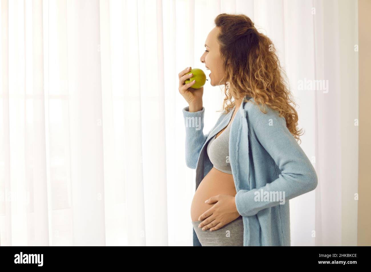 Side view of happy young pregnant lady standing by the window and eating green apple Stock Photo