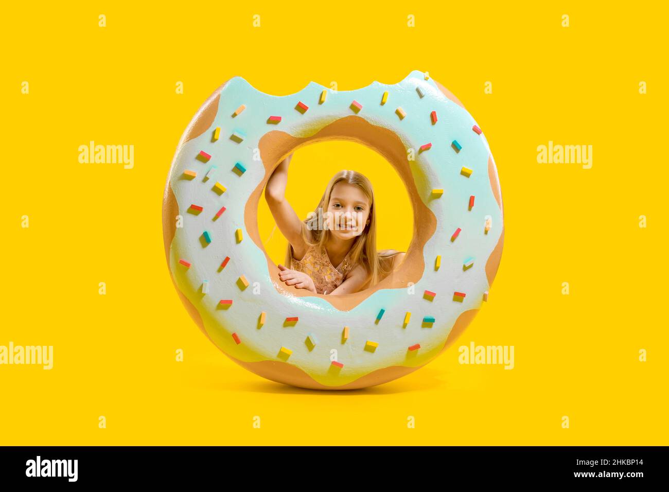 The girl stands behind a huge pink donut with white frosting, looks through the hole. Delicious dessert. Sweetness. Confectionery. Stock Photo