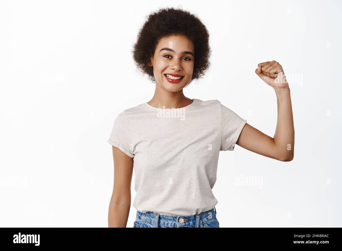 Workout and women power. Smiling african woman showing her strength, flexing biceps, raising arm with muscle, standing over white background Stock Photo
