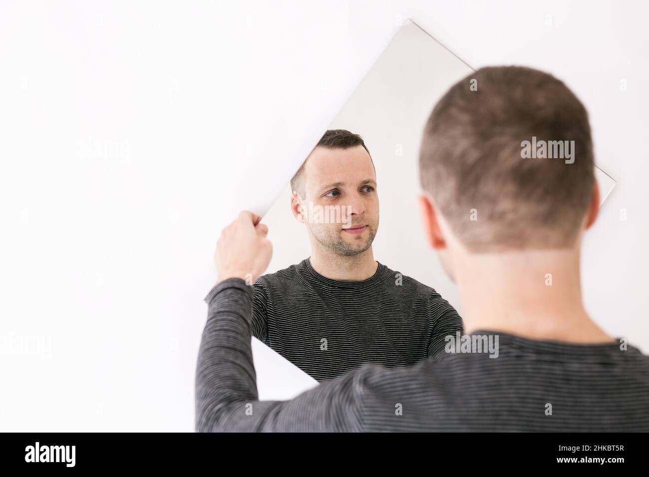Portrait of young man standing in front of a white wall with a mirror in his hands Stock Photo